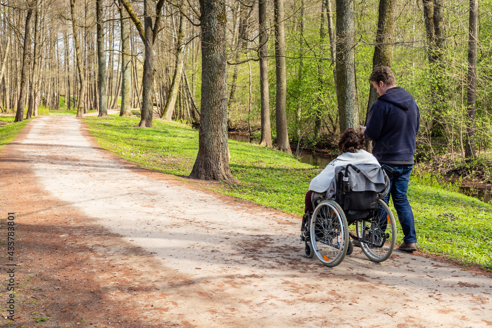 View from back to a woman in wheelchair and a man walking and checking his phone in a park near river at spring. Woman with a disability in wheelchair and her personal assistant are hiking together