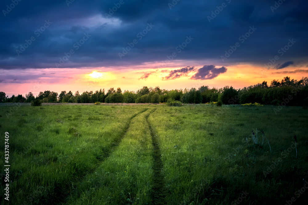 Green field with road and dramatic sky on sunset in summertime.