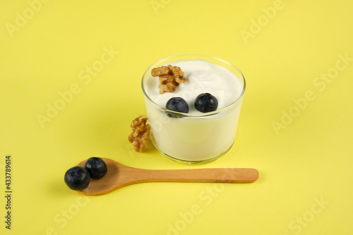 Glass cup with creamy yogurt and blueberries and walnuts with a yellow background and a wooden spoon horizontally