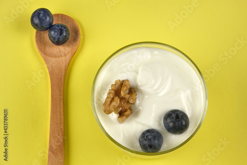 Glass of yogurt with blueberries and walnuts next to small wooden spoon with a yellow background