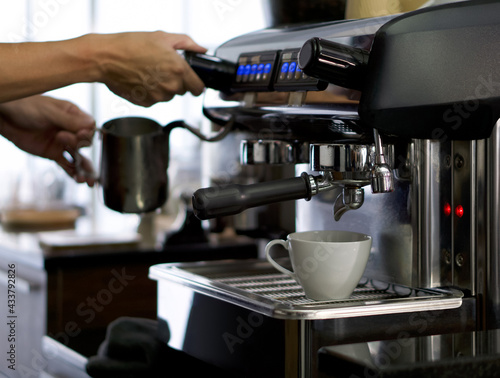 A white ceramic cup placed on the coffee maker. Prepare for making espresso coffee. Barista holding  milk frothing pitcher in the background.