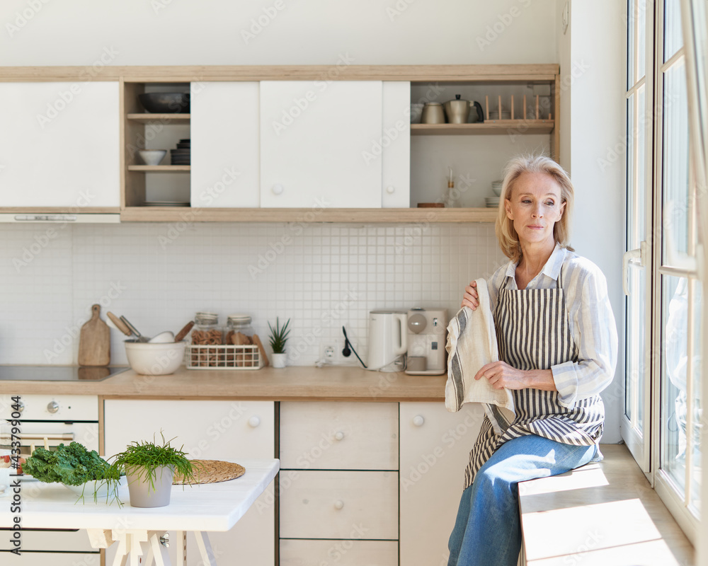 Thoughtful beautiful mature woman looking in window and thinking or dreaming about something, cooking food on kitchen spending free time in morning at home Thoughtful beautiful mature woman looking in window and thinking or dreaming about something, cooking food on kitchen spending free time in morning at home