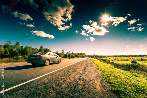 Картината върху платно Car rides along the forest road which is in the field with haystacks lit by the sun on a blue sky background