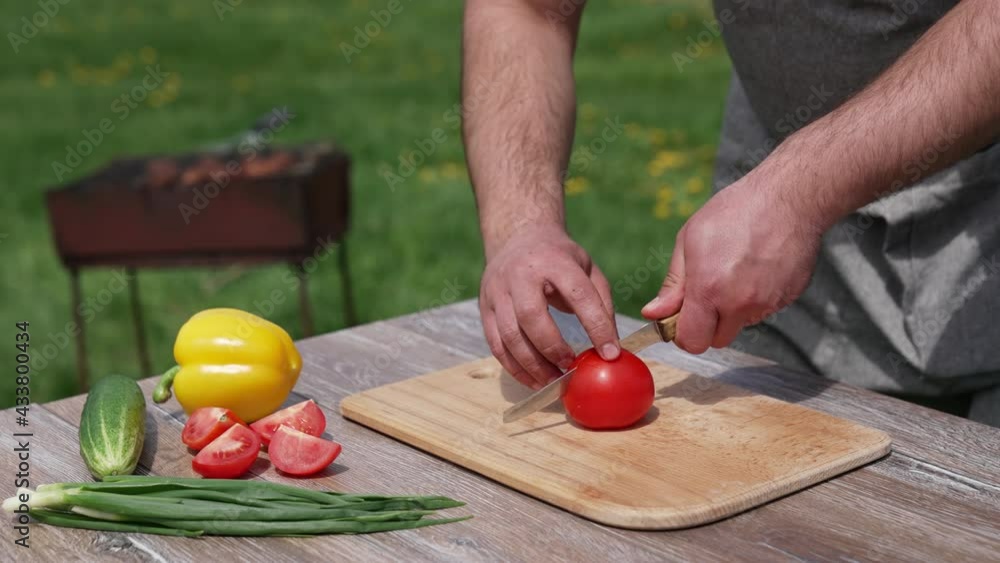Man cutting tomato on a board outdoors. Preparing dinner at dacha. Man cuts vegetable into four parts on a table before the picnic. Man cutting tomato on a board outdoors. Preparing dinner at dacha. Man cuts vegetable into four parts on a table before the picnic.
