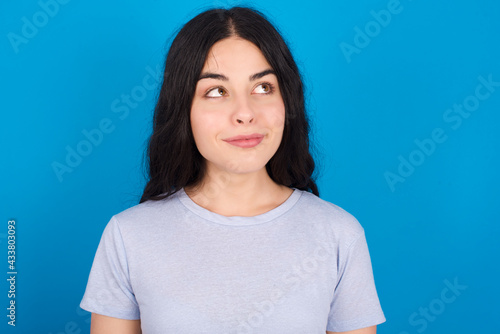 young beautiful tattooed girl wearing blue t-shirt standing against blue background looking aside into empty space thoughtful