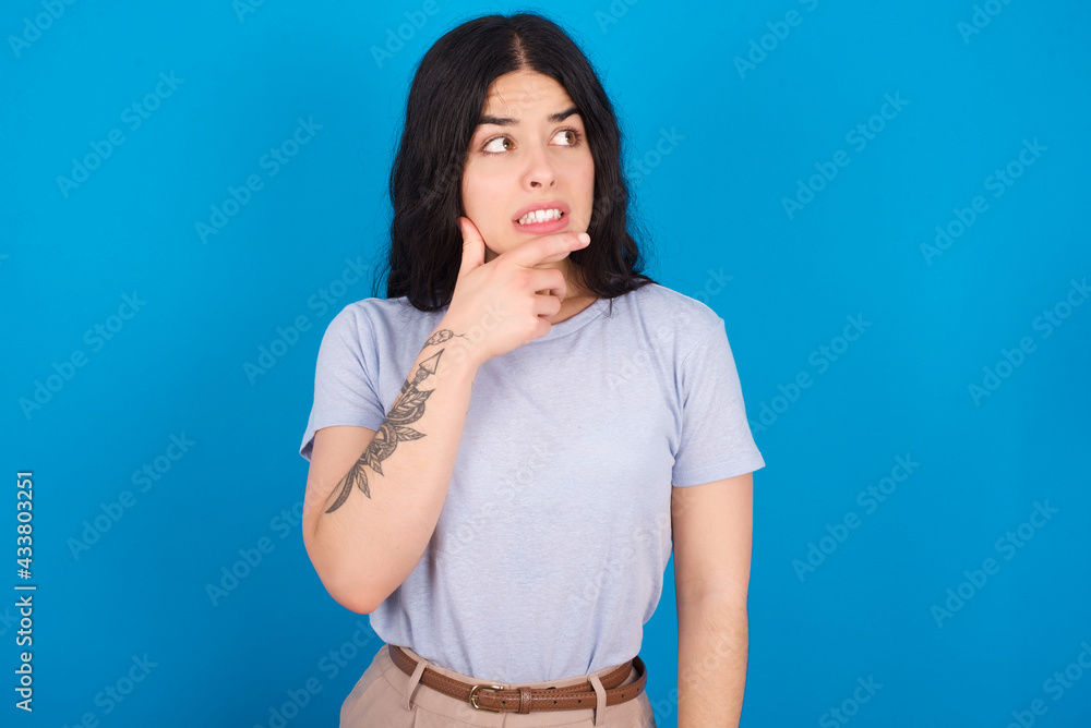 young beautiful tattooed girl wearing blue t-shirt standing against blue background Thinking worried about a question, concerned and nervous with hand on chin. young beautiful tattooed girl wearing blue t-shirt standing against blue background Thinking worried about a question, concerned and nervous with hand on chin.