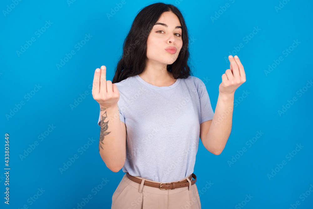 Fototapeta premium young beautiful tattooed girl wearing blue t-shirt standing against blue background doing money gesture with hands, asking for salary payment, millionaire business