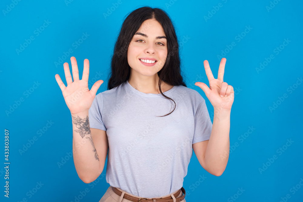 young beautiful tattooed girl wearing blue t-shirt standing against blue background showing and pointing up with fingers number eight while smiling confident and happy. young beautiful tattooed girl wearing blue t-shirt standing against blue background showing and pointing up with fingers number eight while smiling confident and happy.