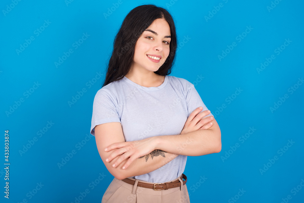 young beautiful tattooed girl wearing blue t-shirt standing against blue background happy face smiling with crossed arms looking at the camera. Positive person. young beautiful tattooed girl wearing blue t-shirt standing against blue background happy face smiling with crossed arms looking at the camera. Positive person.