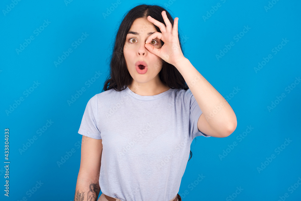 young beautiful tattooed girl wearing blue t-shirt standing against blue background doing ok gesture shocked with surprised face, eye looking through fingers. Unbelieving expression. young beautiful tattooed girl wearing blue t-shirt standing against blue background doing ok gesture shocked with surprised face, eye looking through fingers. Unbelieving expression.