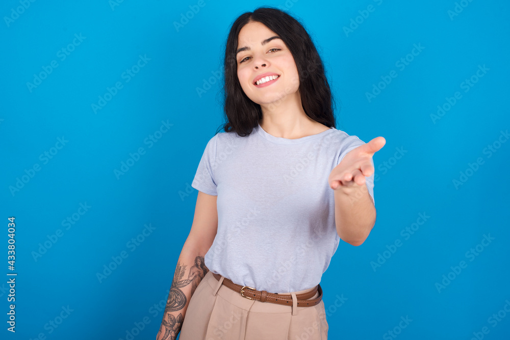 young beautiful tattooed girl wearing blue t-shirt standing against blue background smiling friendly offering handshake as greeting and welcoming. Successful business. young beautiful tattooed girl wearing blue t-shirt standing against blue background smiling friendly offering handshake as greeting and welcoming. Successful business.