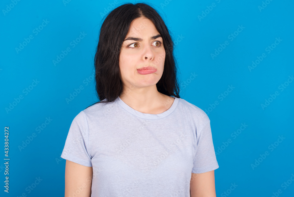 young beautiful tattooed girl wearing blue t-shirt standing against blue background making grimace and crazy face, screaming out of control, funny lunatic expressing freedom and wild. young beautiful tattooed girl wearing blue t-shirt standing against blue background making grimace and crazy face, screaming out of control, funny lunatic expressing freedom and wild.
