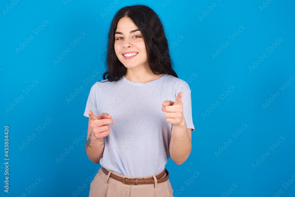 young beautiful tattooed girl wearing blue t-shirt standing against blue background pointing fingers to camera with happy and funny face. Good energy and vibes. young beautiful tattooed girl wearing blue t-shirt standing against blue background pointing fingers to camera with happy and funny face. Good energy and vibes.