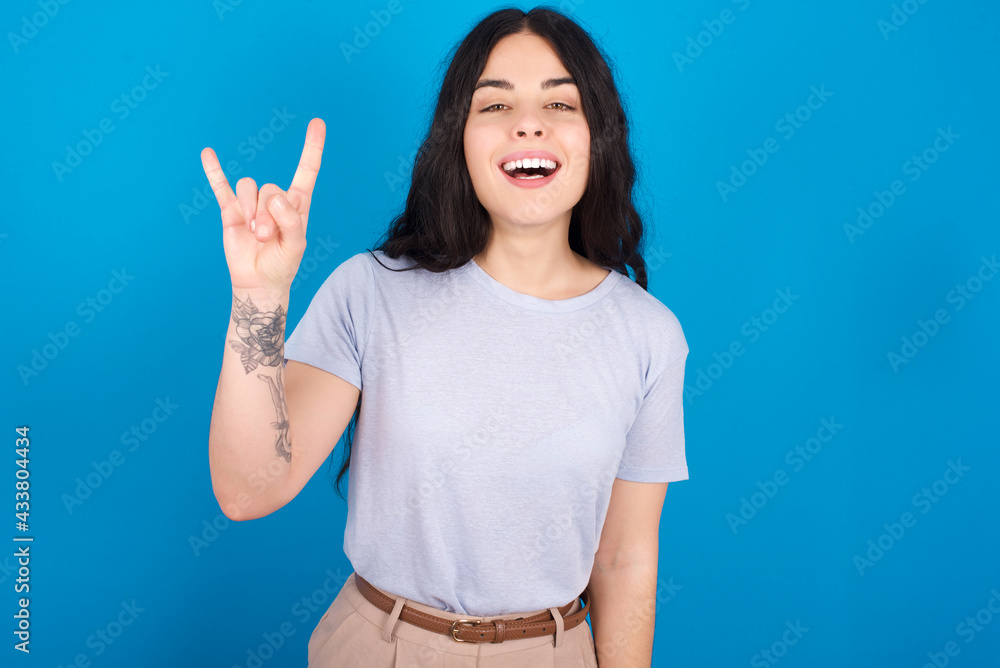young beautiful tattooed girl wearing blue t-shirt standing against blue background doing a rock gesture and smiling to the camera. Ready to go to her favorite band concert. young beautiful tattooed girl wearing blue t-shirt standing against blue background doing a rock gesture and smiling to the camera. Ready to go to her favorite band concert.