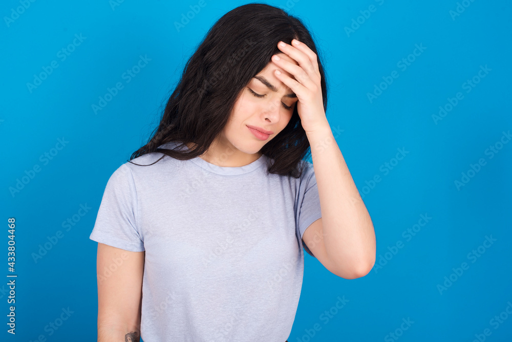 A very upset and lonely young beautiful tattooed girl wearing blue t-shirt standing against blue background crying,