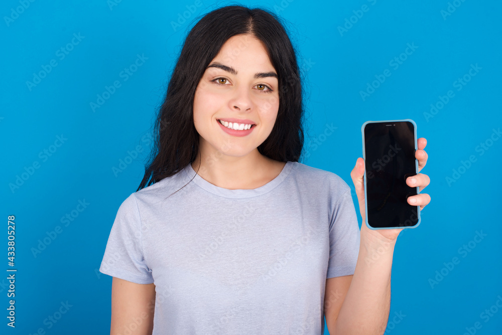 Smiling young beautiful tattooed girl wearing blue t-shirt standing against blue background Mock up copy space. Hold mobile phone with blank empty screen Smiling young beautiful tattooed girl wearing blue t-shirt standing against blue background Mock up copy space. Hold mobile phone with blank empty screen