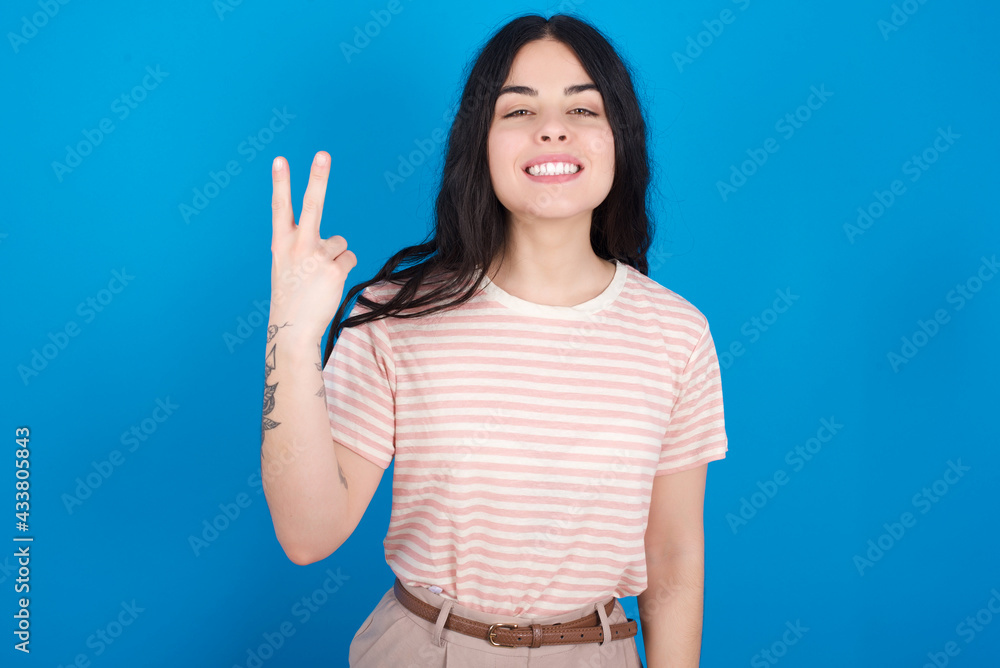 Fototapeta premium young beautiful tattooed girl wearing pink striped t-shirt standing against blue background smiling and looking friendly, showing number two or second with hand forward, counting down