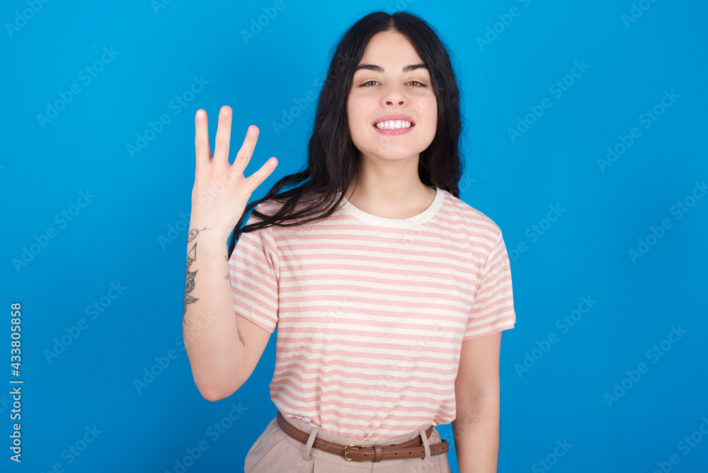 young beautiful tattooed girl wearing pink striped t-shirt standing against blue background smiling and looking friendly, showing number four or fourth with hand forward, counting down young beautiful tattooed girl wearing pink striped t-shirt standing against blue background smiling and looking friendly, showing number four or fourth with hand forward, counting down