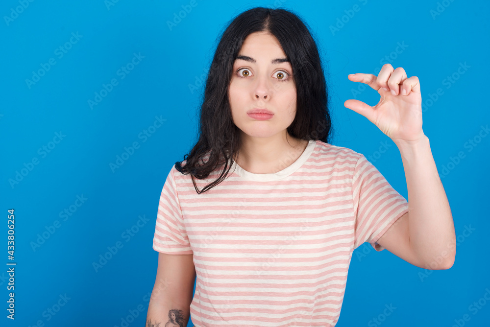 Fototapeta premium young beautiful tattooed girl wearing pink striped t-shirt standing against blue background purses lip and gestures with hand, shows something very little.