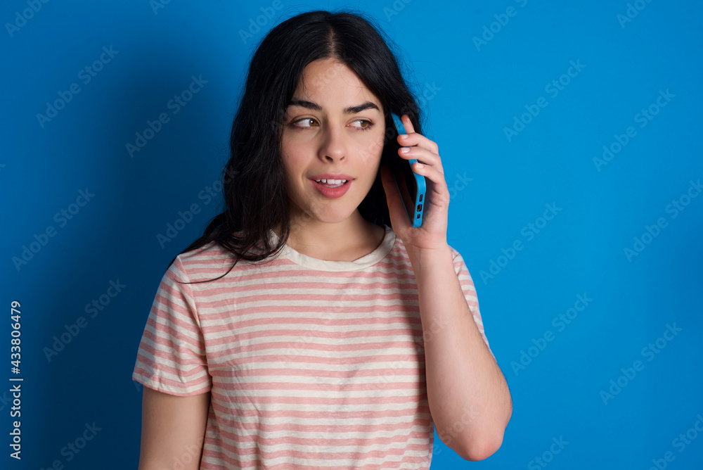 Pleasant looking happy young beautiful tattooed girl wearing pink striped t-shirt has nice telephone conversation and looks aside, has nice mood and smiles positively while talks via cell phone Pleasant looking happy young beautiful tattooed girl wearing pink striped t-shirt has nice telephone conversation and looks aside, has nice mood and smiles positively while talks via cell phone