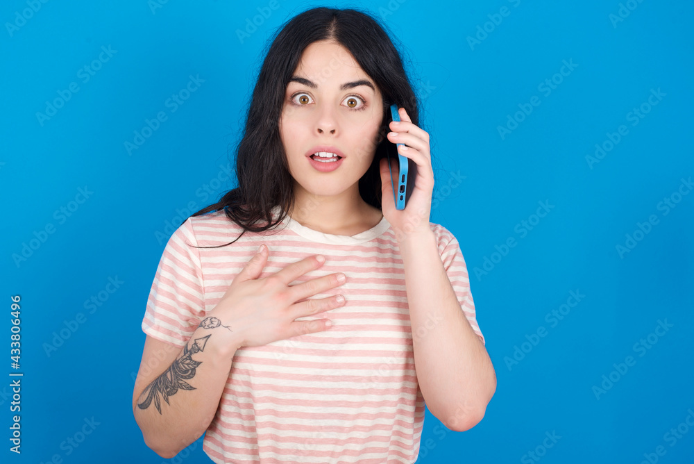 Smiling young beautiful tattooed girl wearing pink striped t-shirt standing against blue background talks via cellphone, enjoys pleasant great conversation. People, technology, communication concept Smiling young beautiful tattooed girl wearing pink striped t-shirt standing against blue background talks via cellphone, enjoys pleasant great conversation. People, technology, communication concept