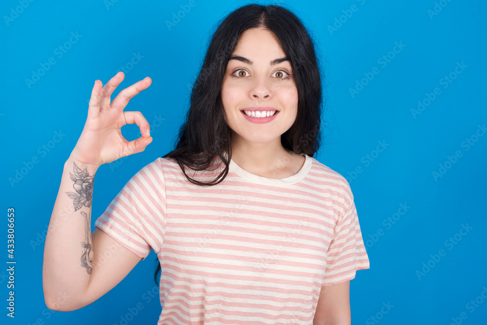 Fototapeta premium young beautiful tattooed girl wearing pink striped t-shirt standing against blue background hold hand arm okey symbol toothy approve advising novelty news