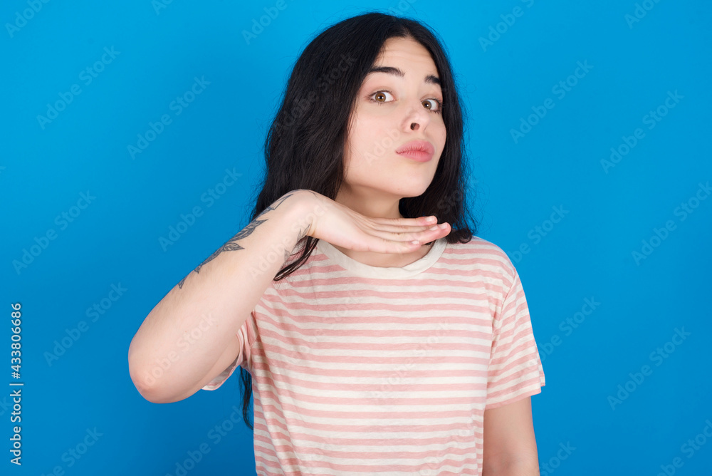 young beautiful tattooed girl wearing pink striped t-shirt standing against blue background cutting throat with hand as knife, threaten aggression with furious violence. young beautiful tattooed girl wearing pink striped t-shirt standing against blue background cutting throat with hand as knife, threaten aggression with furious violence.