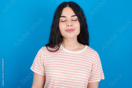 young beautiful tattooed girl wearing pink striped t-shirt standing against blue background nice-looking sweet charming cute attractive lovely winsome sweet peaceful closed eyes
