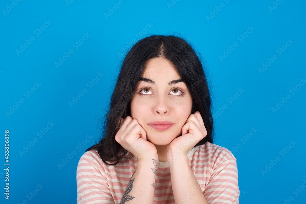 Portrait of sad young beautiful tattooed girl wearing pink striped t-shirt standing against blue background hands face look empty space Portrait of sad young beautiful tattooed girl wearing pink striped t-shirt standing against blue background hands face look empty space