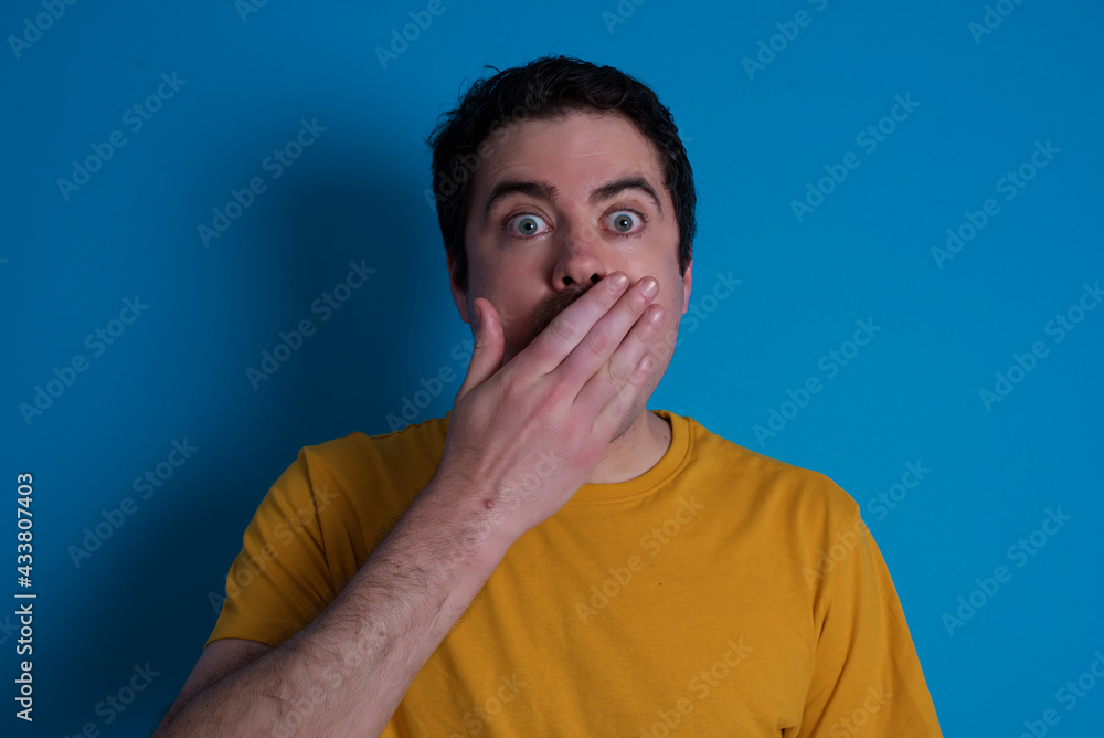 Amazed young handsome Caucasian man with moustache wearing orange t-shirt against blue background bitting lip and looking tricky to empty space. Amazed young handsome Caucasian man with moustache wearing orange t-shirt against blue background bitting lip and looking tricky to empty space.