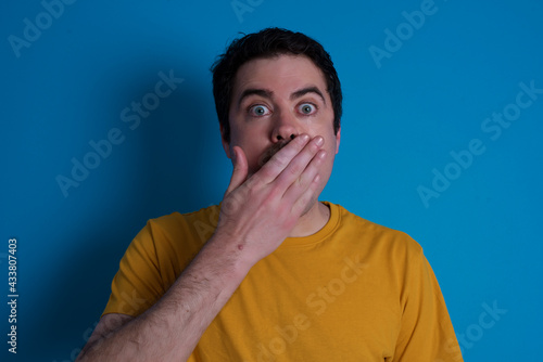 Amazed young handsome Caucasian man with moustache wearing orange t-shirt against blue background bitting lip and looking tricky to empty space.