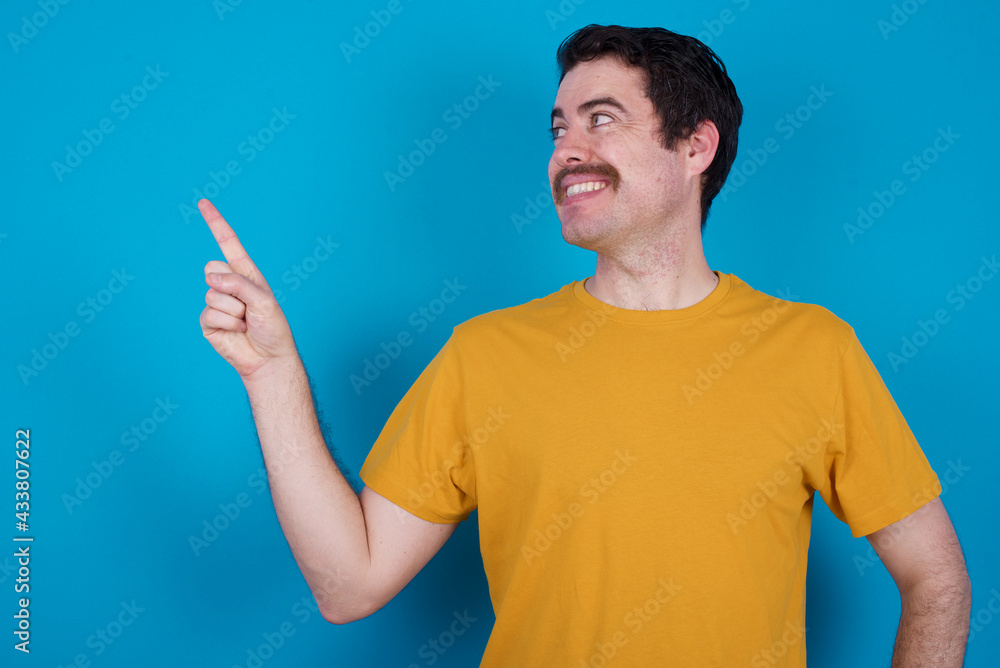 Smiling young handsome Caucasian man with moustache wearing orange t-shirt against blue background indicating finger empty space showing best low prices Smiling young handsome Caucasian man with moustache wearing orange t-shirt against blue background indicating finger empty space showing best low prices