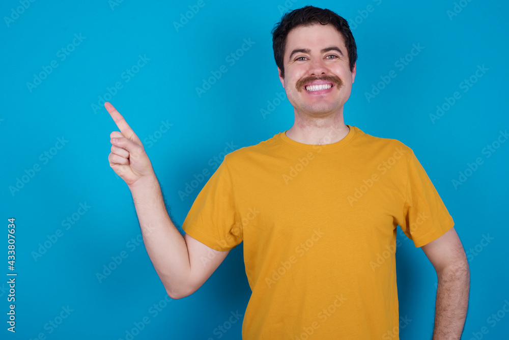 Fototapeta premium young handsome Caucasian man with moustache wearing orange t-shirt against blue background looking at camera indicating finger empty space sales