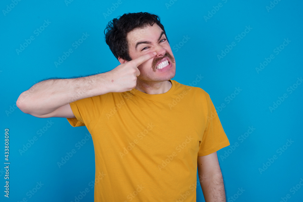 Φωτογραφία young handsome Caucasian man with moustache wearing orange t-shirt against blue background pointing unhappy to pimple on forehead, ugly infection of blackhead