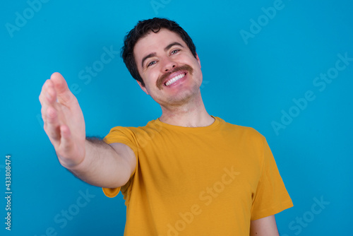 young handsome Caucasian man with moustache wearing yellow t-shirt against blue background smiling friendly offering handshake as greeting and welcoming. Successful business.