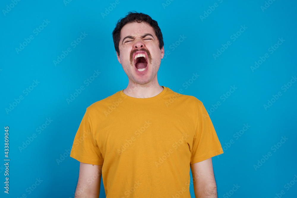 young handsome Caucasian man with moustache wearing yellow t-shirt against blue background angry and mad screaming frustrated and furious, shouting with anger. Rage and aggressive concept. young handsome Caucasian man with moustache wearing yellow t-shirt against blue background angry and mad screaming frustrated and furious, shouting with anger. Rage and aggressive concept.