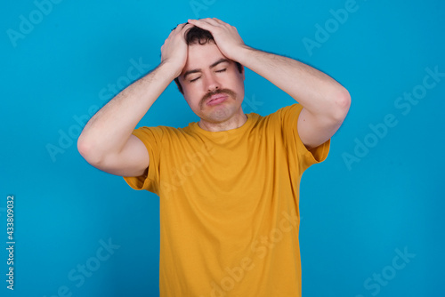 young handsome Caucasian man with moustache wearing yellow t-shirt against blue background suffering from strong headache desperate and stressed because of overwork. Depression and pain concept.
