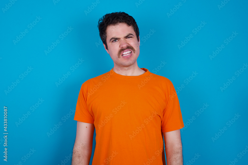 Portrait of dissatisfied young handsome Caucasian man with moustache wearing orange t-shirt against smirks face, purses lips and looks with annoyance at camera, discontent hearing something unpleasant Portrait of dissatisfied young handsome Caucasian man with moustache wearing orange t-shirt against smirks face, purses lips and looks with annoyance at camera, discontent hearing something unpleasant