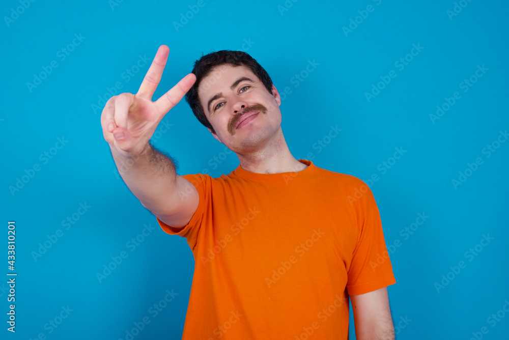 young handsome Caucasian man with moustache wearing orange t-shirt against blue background directs fingers at camera selects someone. I recommend you. Best choice young handsome Caucasian man with moustache wearing orange t-shirt against blue background directs fingers at camera selects someone. I recommend you. Best choice