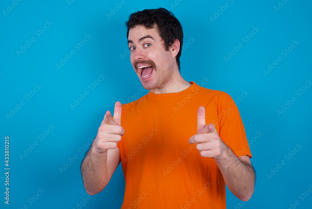 young handsome Caucasian man with moustache wearing orange t-shirt against blue background directs fingers at camera selects someone. I recommend you. Best choice young handsome Caucasian man with moustache wearing orange t-shirt against blue background directs fingers at camera selects someone. I recommend you. Best choice
