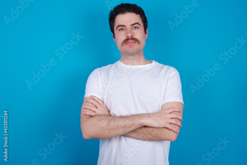 Confident young handsome Caucasian man with moustache wearing white t-shirt against white background with arms crossed looking to the camera