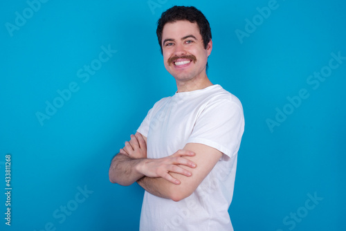 Portrait of young handsome Caucasian man with moustache wearing white t-shirt against white background standing with folded arms and smiling
