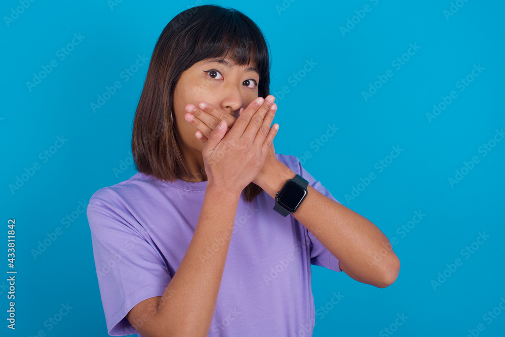 Young beautiful asian girl wearing purple t-shirt against blue background shocked covering mouth with hands for mistake. Secret concept. Young beautiful asian girl wearing purple t-shirt against blue background shocked covering mouth with hands for mistake. Secret concept.