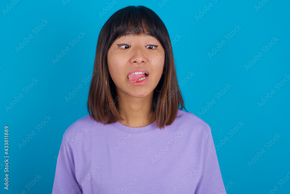 Young beautiful asian girl wearing purple t-shirt against blue background showing grimace face crossing eyes and showing tongue. Being funny and crazy Young beautiful asian girl wearing purple t-shirt against blue background showing grimace face crossing eyes and showing tongue. Being funny and crazy