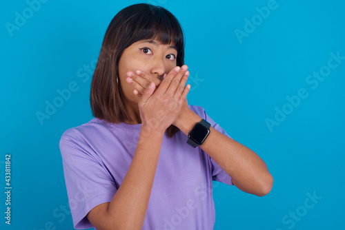 Young beautiful asian girl wearing purple t-shirt against blue background shocked covering mouth with hands for mistake. Secret concept.