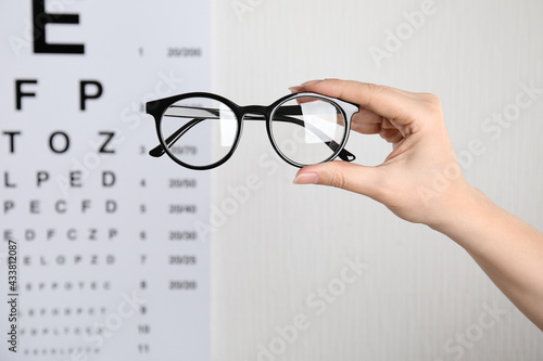 Woman holding glasses against eye chart on light background, closeup. Ophthalmologist prescription