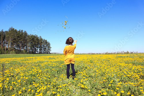 A ten-year-old child in a yellow dress throws a bouquet of dandelions up in a spring meadow against a blue sky