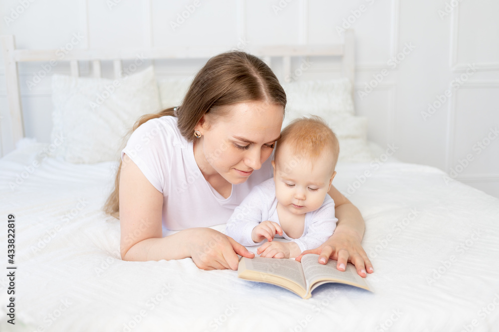 mom reads a book to baby before going to bed on a white bed with cotton bedding at home, happy family mom reads a book to baby before going to bed on a white bed with cotton bedding at home, happy family