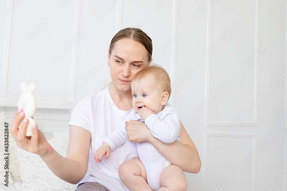 a happy mother holds a baby in her arms showing a teether or a pear and hugging it on the bed at home a happy mother holds a baby in her arms showing a teether or a pear and hugging it on the bed at home