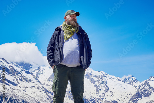 A man with a beard in tourist clothes poses against the backdrop of mountains covered with snow. Travel and adventure.
